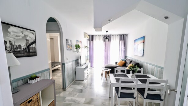 Una sala de estar moderna con comedor, que incluye un sofá, una mesa de centro y una mesa de comedor con sillas. Hay un pasillo que conduce a otra habitación, y las paredes están decoradas con cuadros enmarcados.