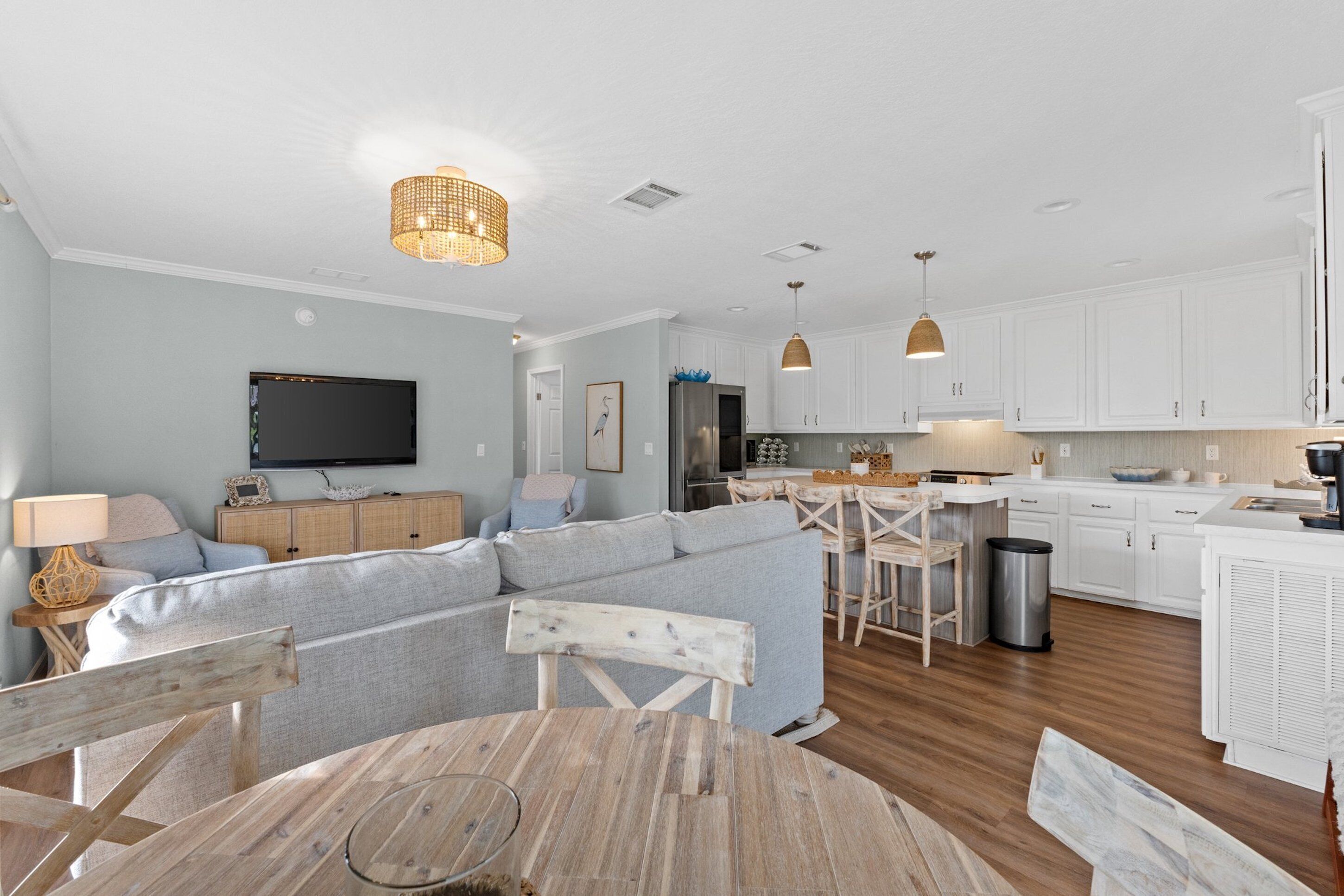 A modern kitchen with white cabinets, a dining area with a round wooden table, and a living room with a grey sofa and a television.