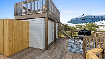 Una terraza de madera con un área de estar techada al aire libre, una parrilla para barbacoa y una escalera que sube a un nivel superior.
