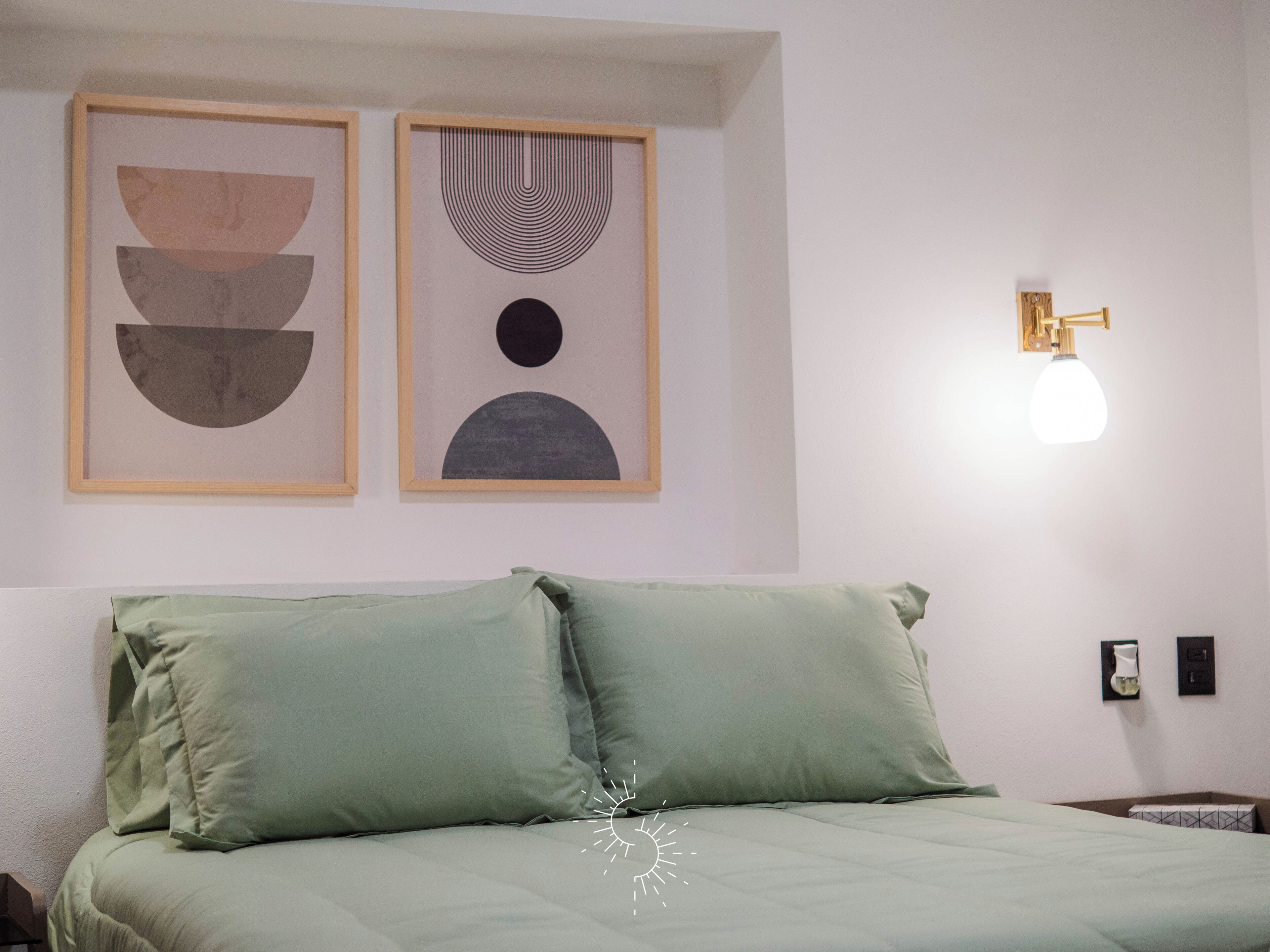 Un dormitorio con una cama, dos obras de arte abstractas enmarcadas, una lámpara de pared y una mesita de noche con lámpara.