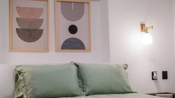 Un dormitorio con una cama, dos obras de arte abstractas enmarcadas, una lámpara de pared y una mesita de noche con lámpara.