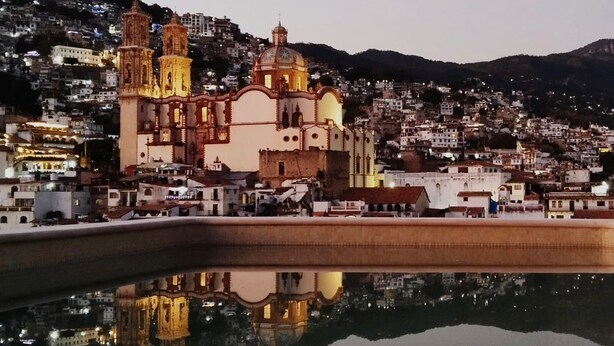 A historic church with a prominent bell tower and dome, reflecting in a calm body of water, surrounded by a densely populated hillside town. - Hotel Casa Domínguez (Taxco)