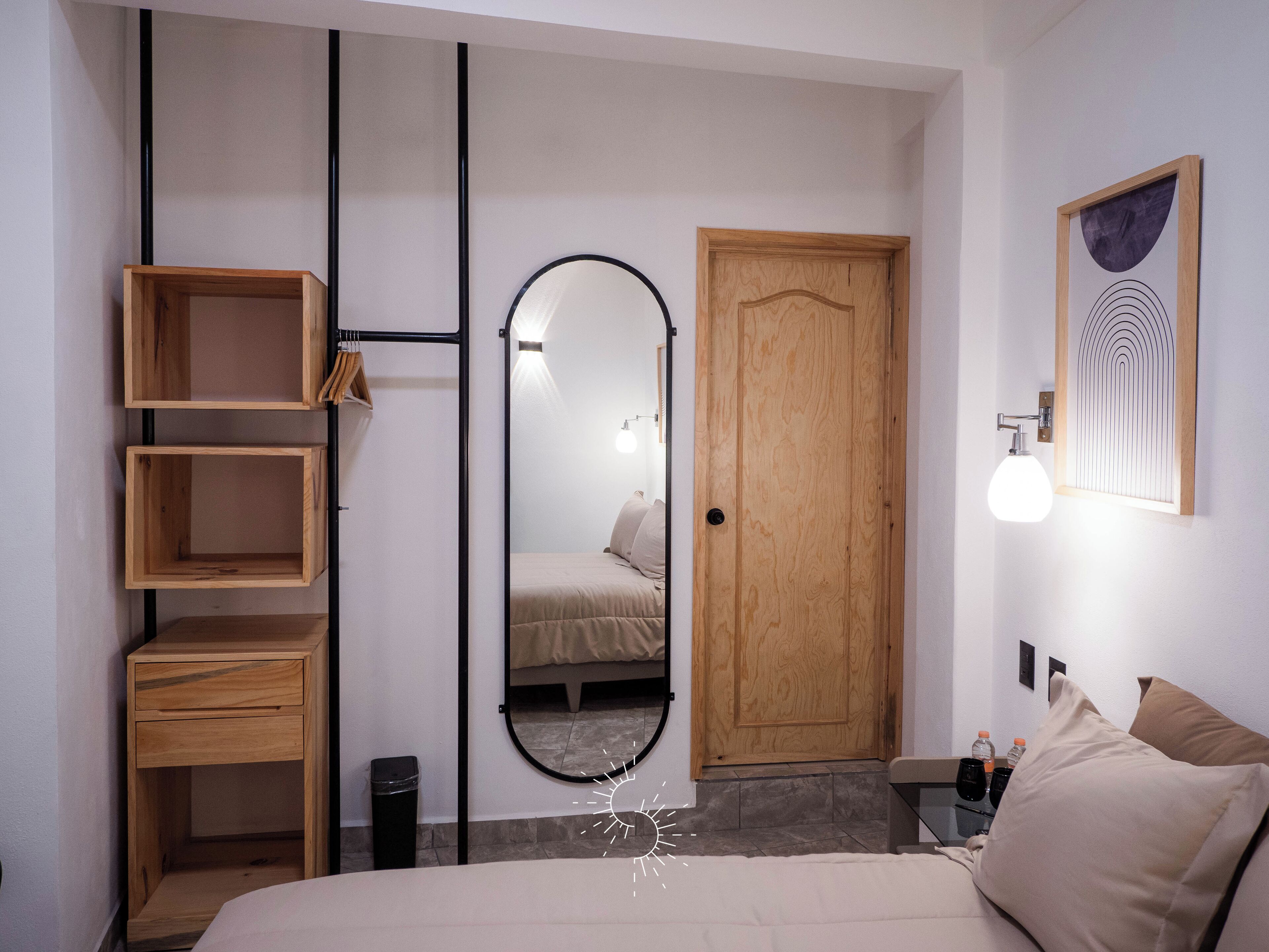 A bedroom with a bed, a wooden wardrobe, a mirror, a door, and a wall-mounted light fixture.