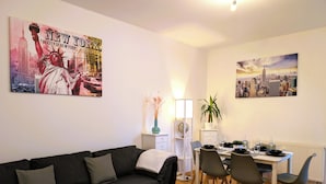 A modern living room with a black sofa, a dining table with chairs, and two cityscape paintings on the walls. - CaSa Apartment East Field - 5 beds, family-friendly, fully equipped (Magdeburg)