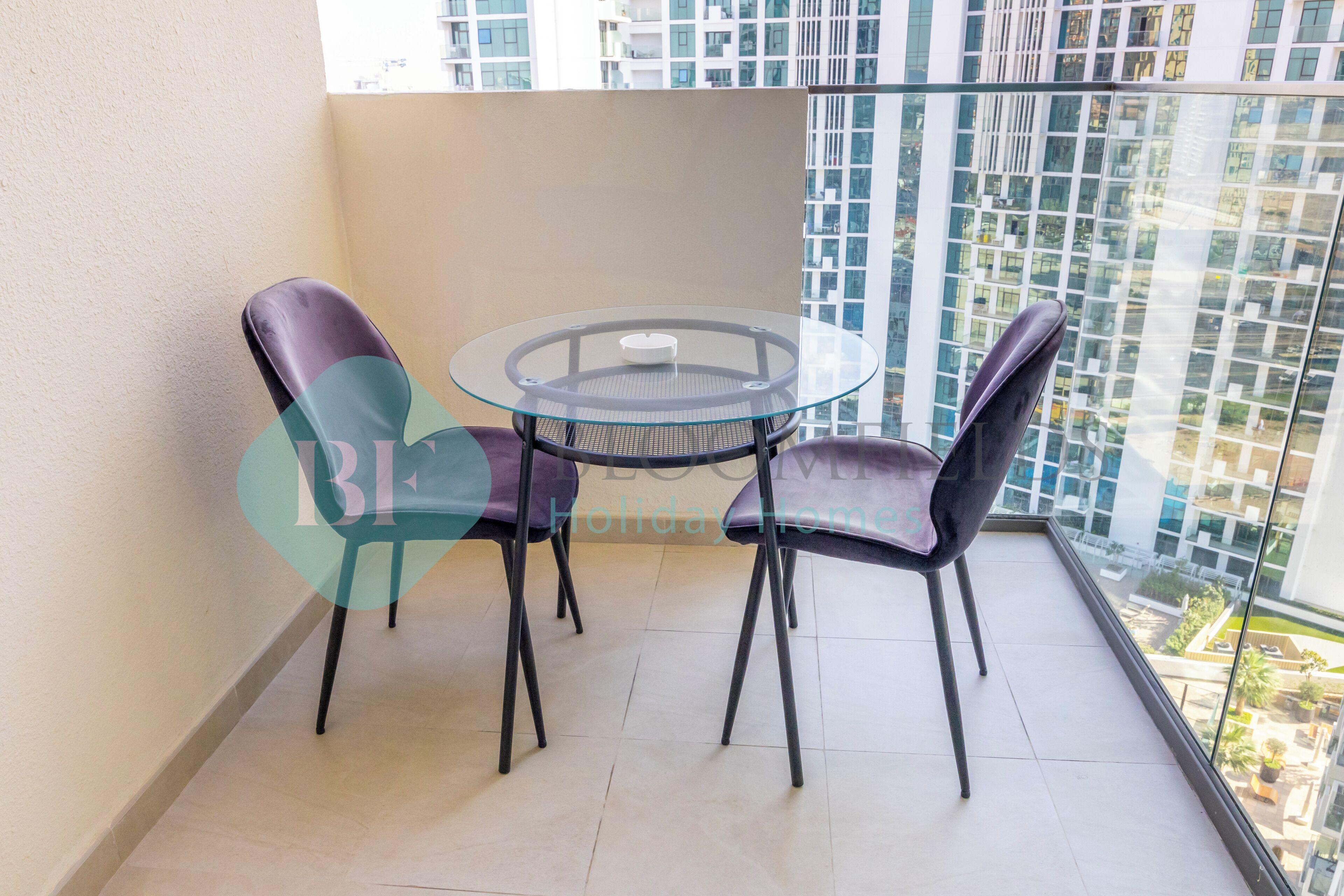 A balcony with a glass table and two chairs, overlooking a cityscape.