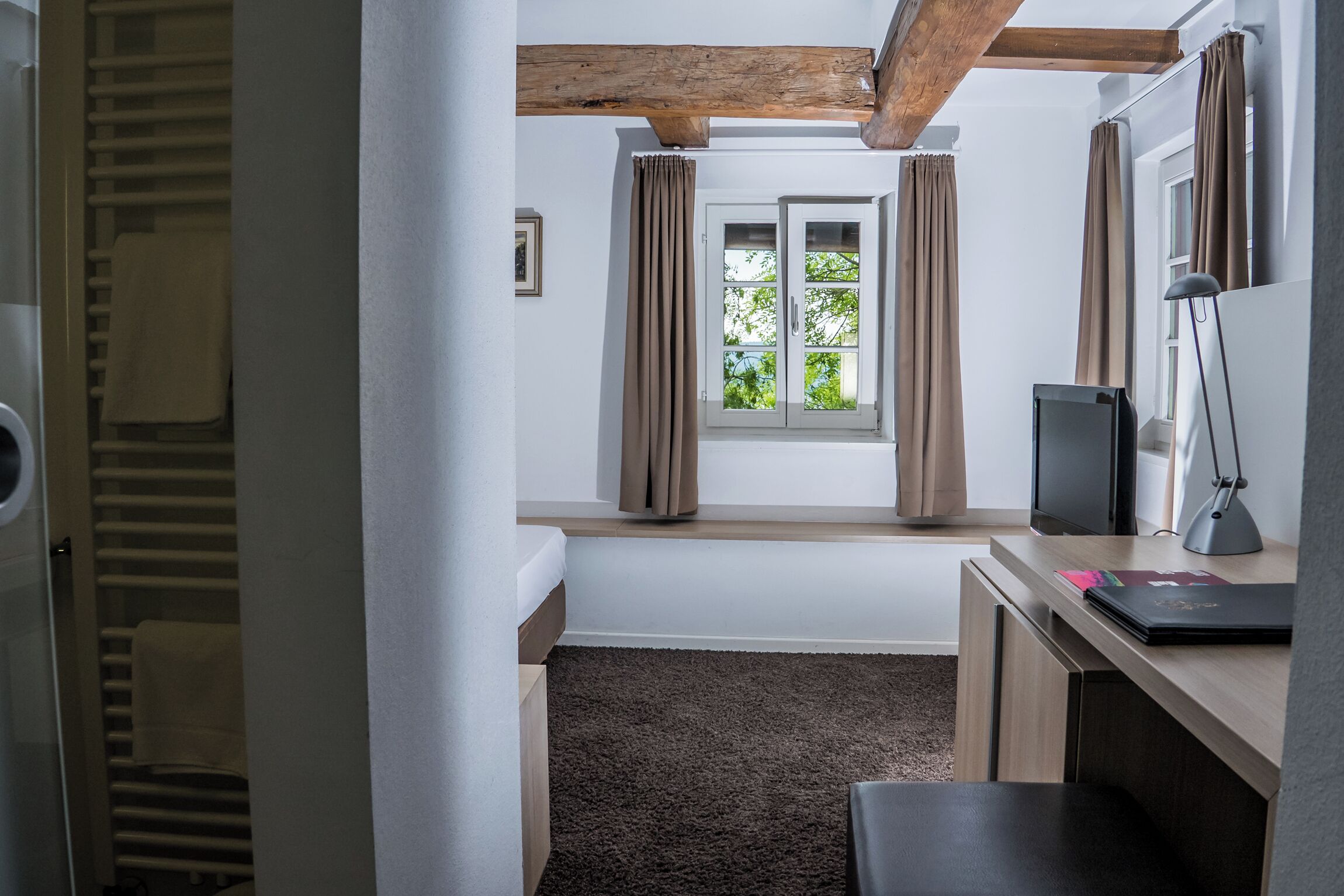 Ein Zimmer mit Holzbalkendecke, einem fenster mit Vorhängen, einem Schreibtisch mit Computer und Lampe sowie einem Teppichboden.
