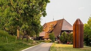 A traditional building with a steeply pitched roof, surrounded by lush greenery and a clear road. - Hotel Hofgut Hohenkarpfen (Hausen ob Verena)