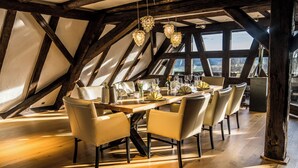 A dining area with a wooden table set for six, surrounded by six chairs, in a room with exposed wooden beams and large windows. - Hotel Hofgut Hohenkarpfen (Hausen ob Verena)