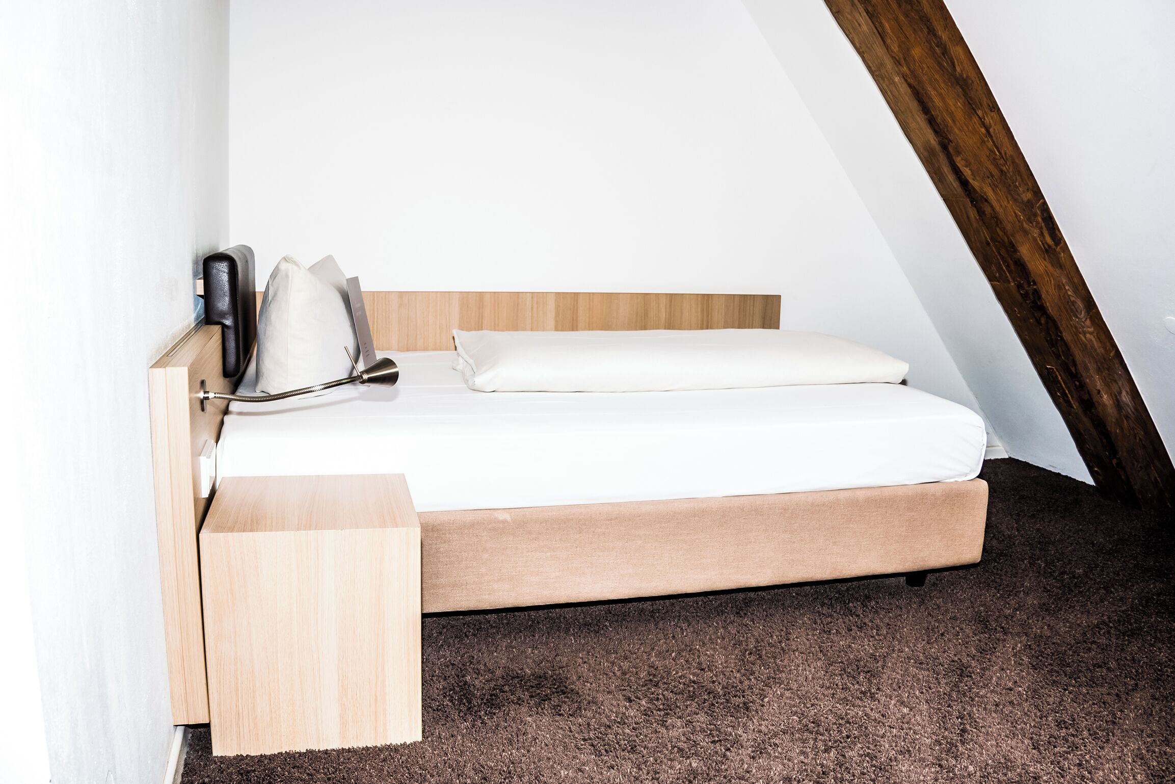 A single bed with a wooden frame and white bedding in a room with sloped ceilings and a brown carpet.