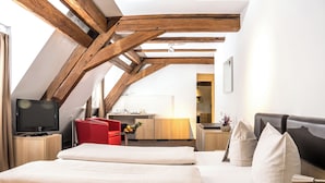 A modern hotel room with a sloped ceiling, wooden beams, a bed with white linens, a red chair, a desk with a lamp, and a television. - Hotel Hofgut Hohenkarpfen (Hausen ob Verena)