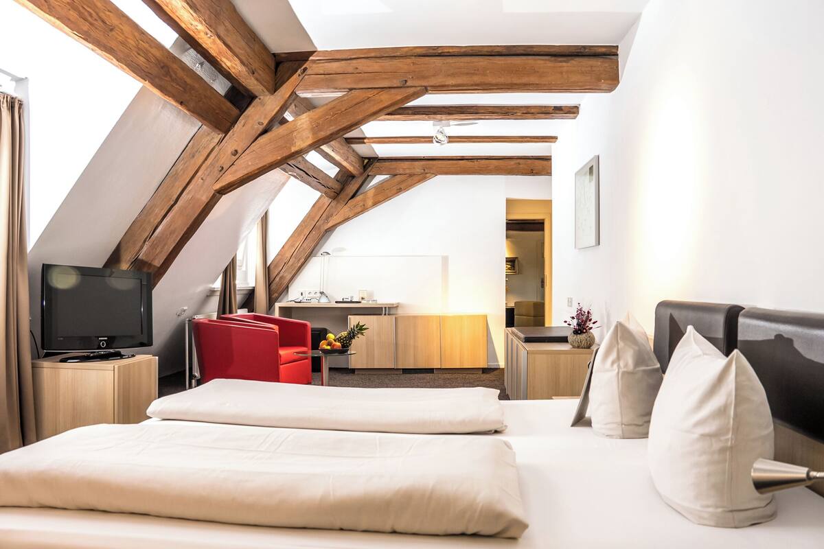 Une chambre d’hôtel moderne avec un plafond en pente, des poutres en bois, un lit avec du linge de maison blanc, une chaise rouge, un bureau avec une lampe et un téléviseur.
