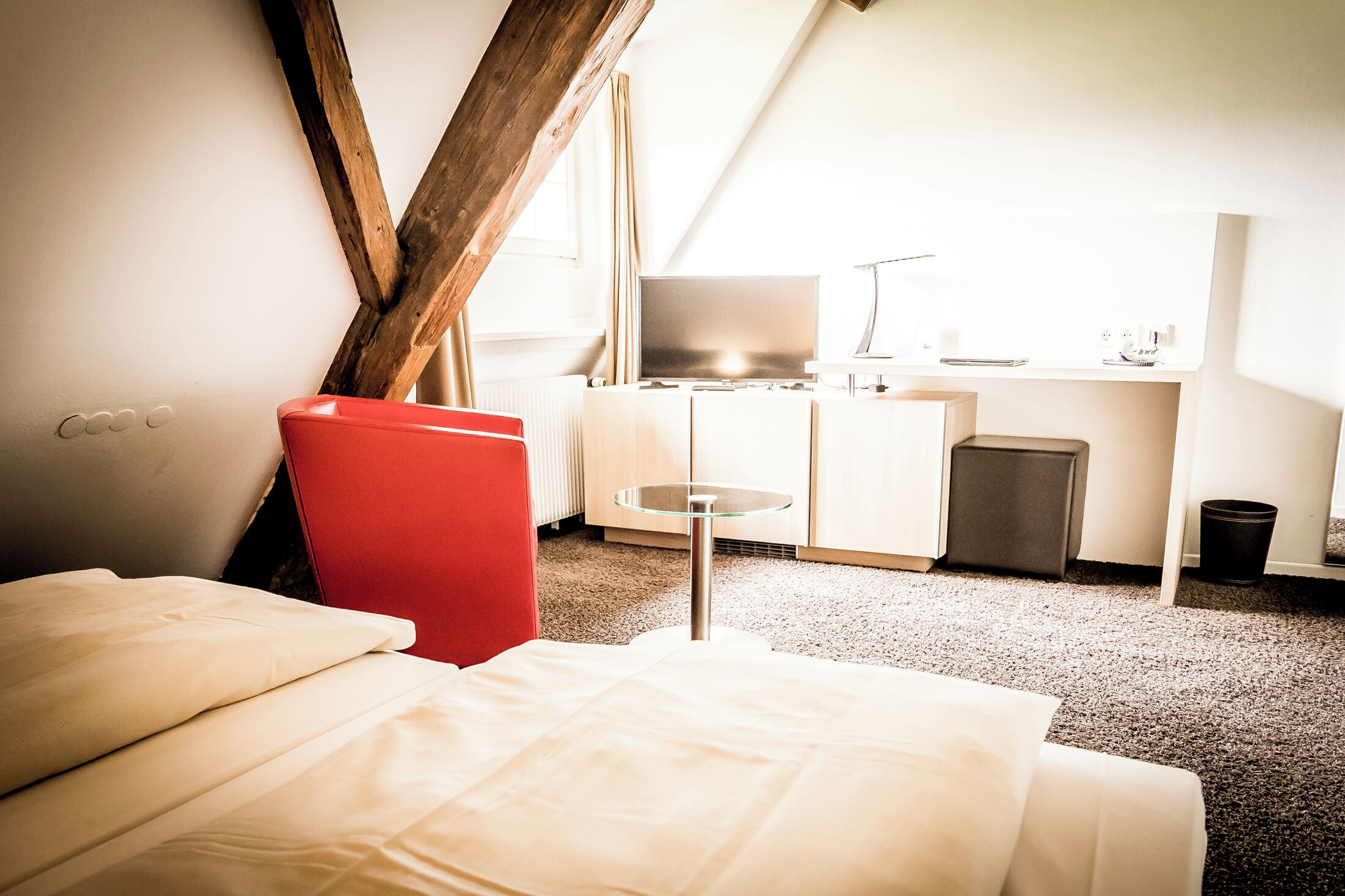Una habitación de hotel con una cama, una silla roja, una mesita, un escritorio con una computadora y un televisor.
