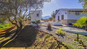 Une maison blanche avec une petite dépendance attenante, entourée d’un jardin où poussent des cactus et des palmiers, et où est installé un hamac.