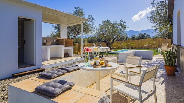 Une terrasse extérieure avec une table, des chaises et des coussins. On aperçoit une piscine au fond et une vue sur des arbres et des montagnes.