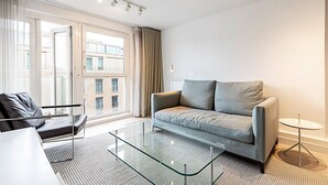 Ett modernt vardagsrum med en grå soffa, ett soffbord i glas och utsikt över en byggnad genom stora fönster.