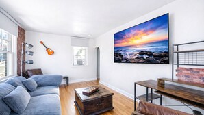 Een woonkamer met een groot raam, een gitaar, een houten salontafel, een bank en een schilderij van een zonsondergang boven de oceaan.
