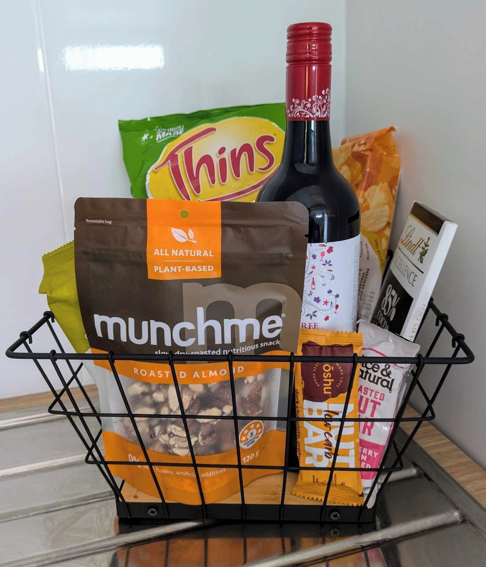 A shopping basket containing a bottle of wine, a bag of roasted almonds, a packet of munchme snacks, a pack of Thins potato chips, and a bar snack package.