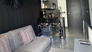 Uma sala de estar moderna com um sofá cinzento, cadeiras pretas, um ventilador de teto e uma área de cozinha ao fundo.