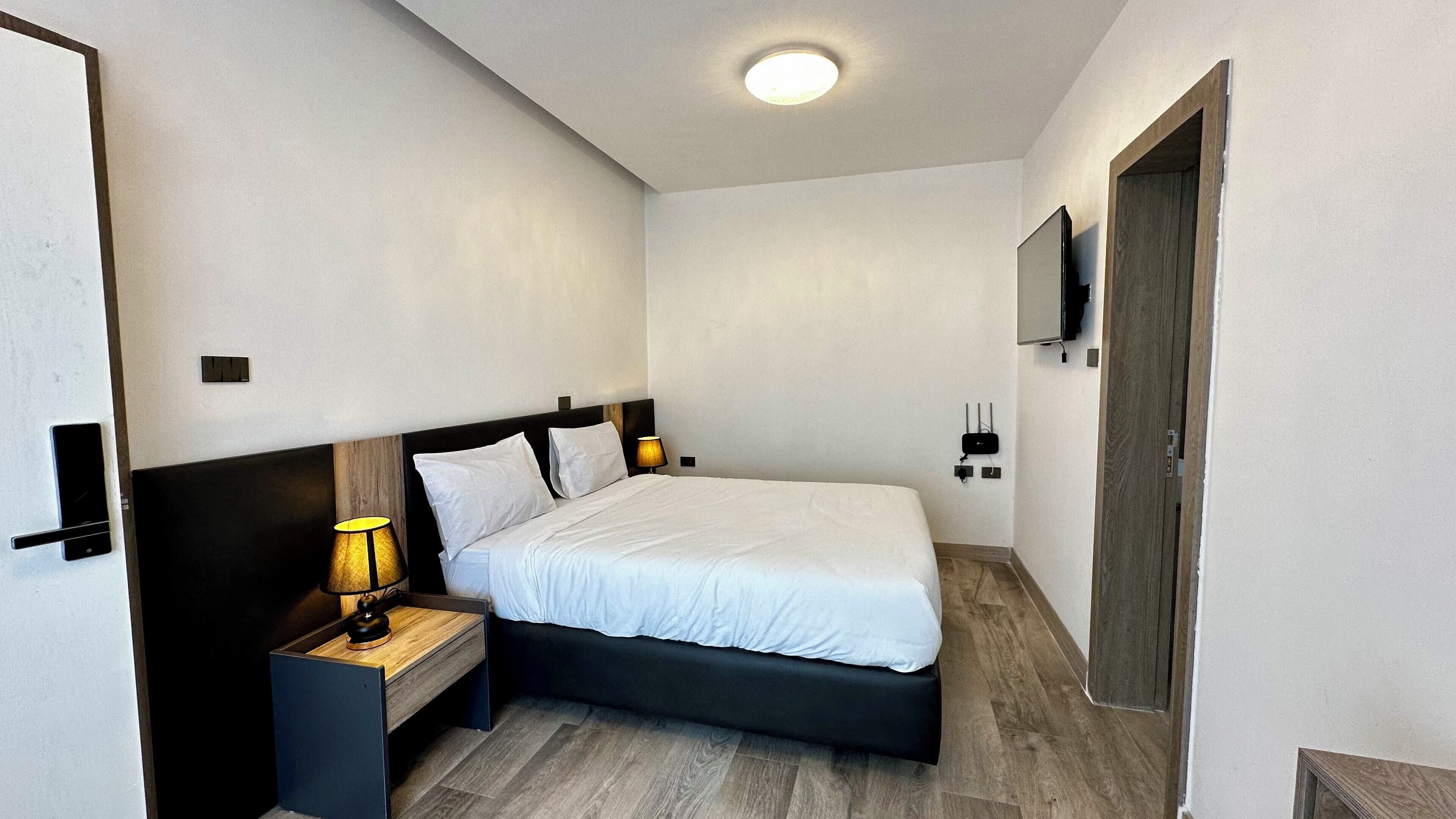 A modern hotel room with a single bed, bedside tables, a lamp, a flat-screen TV mounted on the wall, and a wooden door.