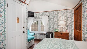 Un dormitorio con papel tapiz floral, una cama con colcha estampada, un escritorio con lámpara y una ventana con cortinas.