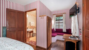 Une pièce avec un papier peint à rayures, une porte en bois, un lit recouvert d’une couvre-lit à motifs floraux, un canapé violet, une petite table avec un livre et un téléviseur fixé au mur.