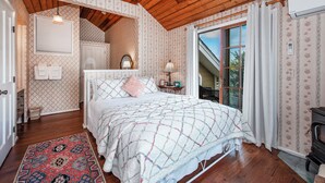 Une chambre avec un plafond en bois, un grand lit recouvert d’une courtepointe, une table de chevet, une commode et une fenêtre avec des rideaux.