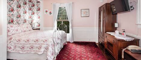 A bedroom with a floral-patterned headboard, a red patterned carpet, a wooden dresser, a bedside table with a lamp, and a television mounted on the wall.