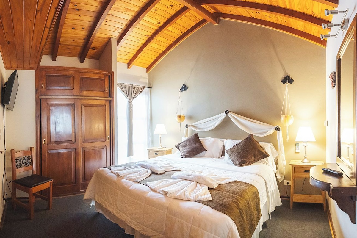 A bedroom with a large bed, wooden ceiling beams, a wardrobe, bedside tables with lamps, and a mirror.