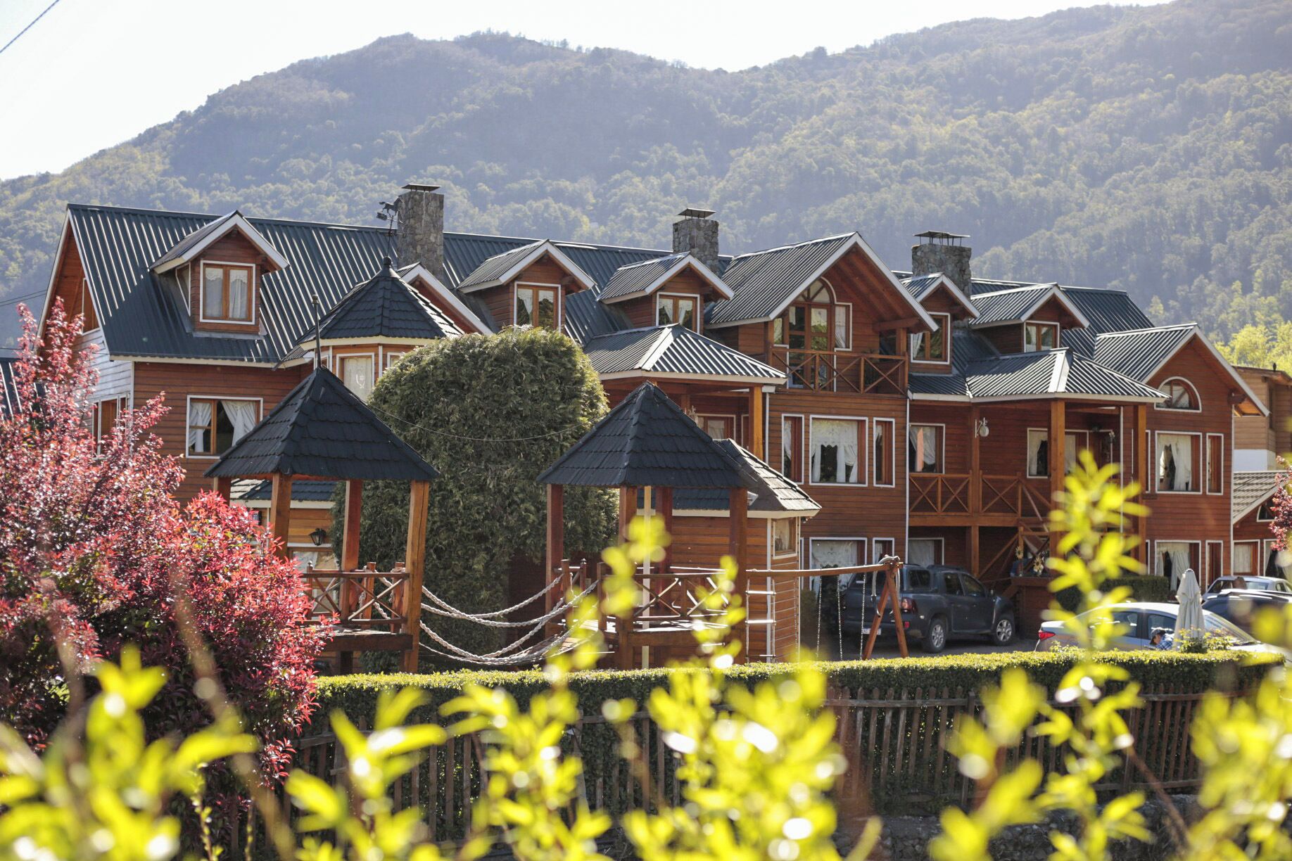A large wooden building with multiple roofs and chimneys, surrounded by a well-maintained hedge and trees.