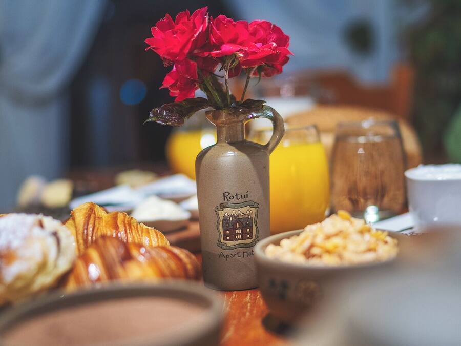 A breakfast table with pastries, a bottle of coffee, and a vase with red flowers.