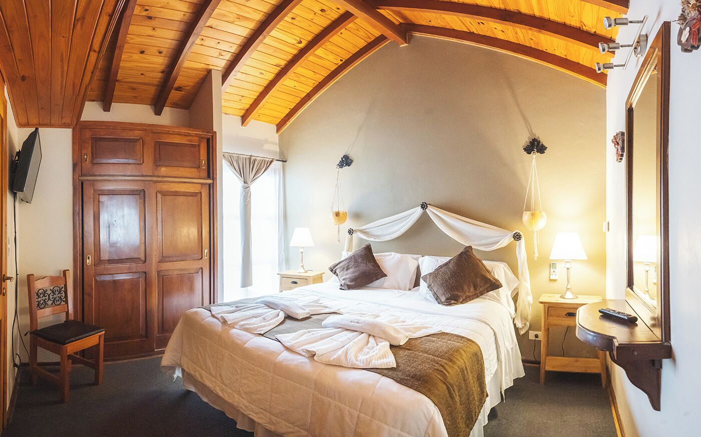 Un dormitorio con una cama grande, vigas de madera en el techo, un armario, mesitas de noche con lámparas y un espejo.