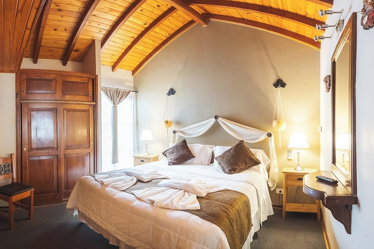 Une chambre à coucher avec un grand lit, des poutres apparentes en bois au plafond, une armoire, des tables de chevet avec des lampes et un miroir.