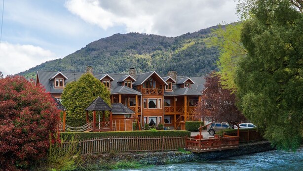 A large wooden lodge with multiple balconies, situated by a river with a mountain backdrop. - Rotui Apart Hotel (San Martín de los Andes)