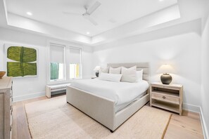 Relax in this serene bedroom with a large bed, nightstand, and bench, surrounded by a ceiling fan and a beautiful artwork.