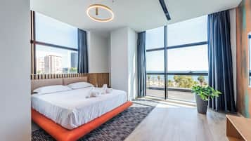 A modern hotel room with a large bed, a view of the city and sea, and a potted plant.