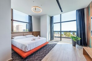 Ein modernes Hotelzimmer mit einem großen Bett, Blick auf Stadt und Meer sowie einer Zimmerpflanze.