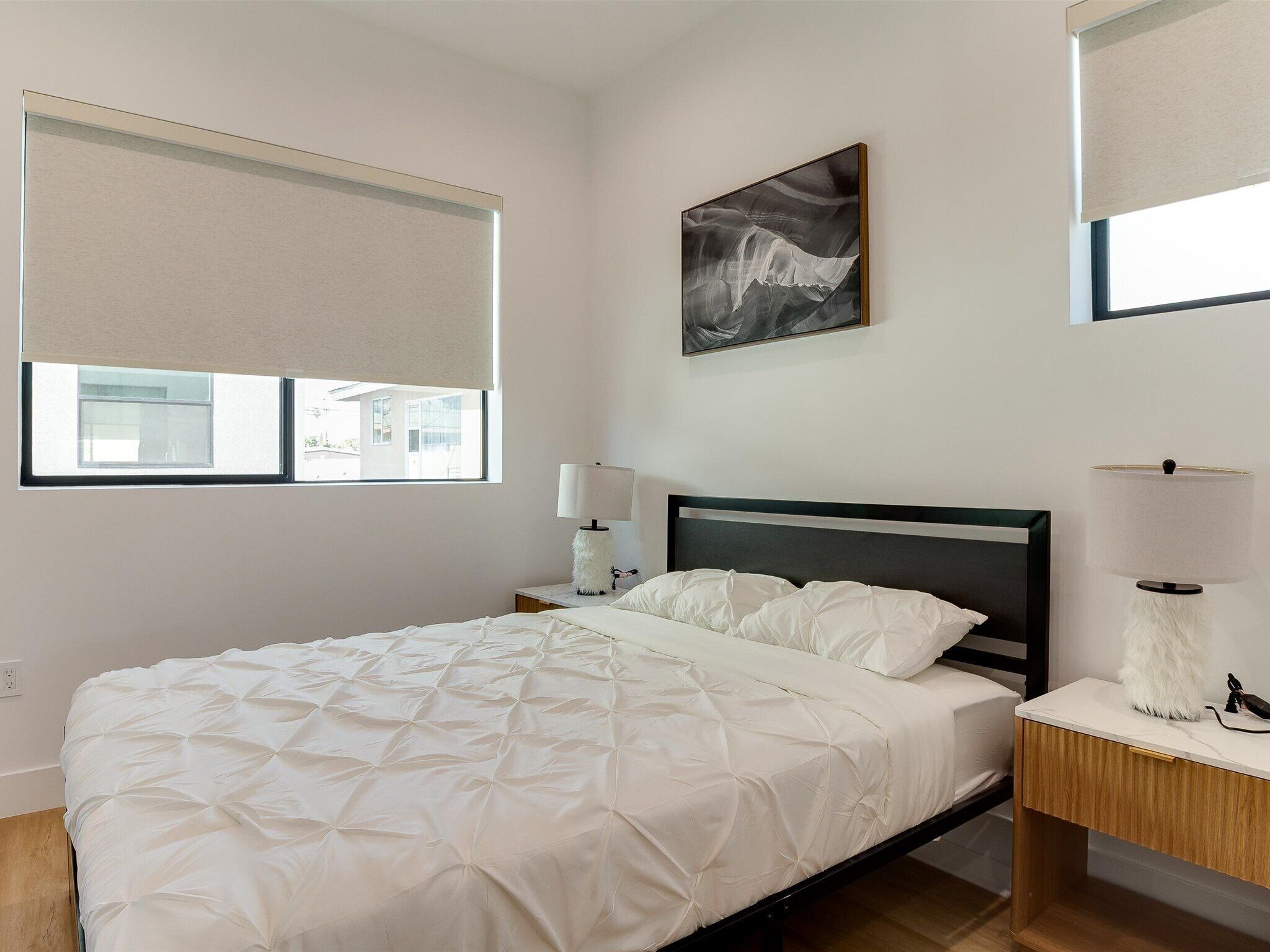 Un dormitorio con cama, mesita de noche, lámpara, cuadro y ventana con persianas.