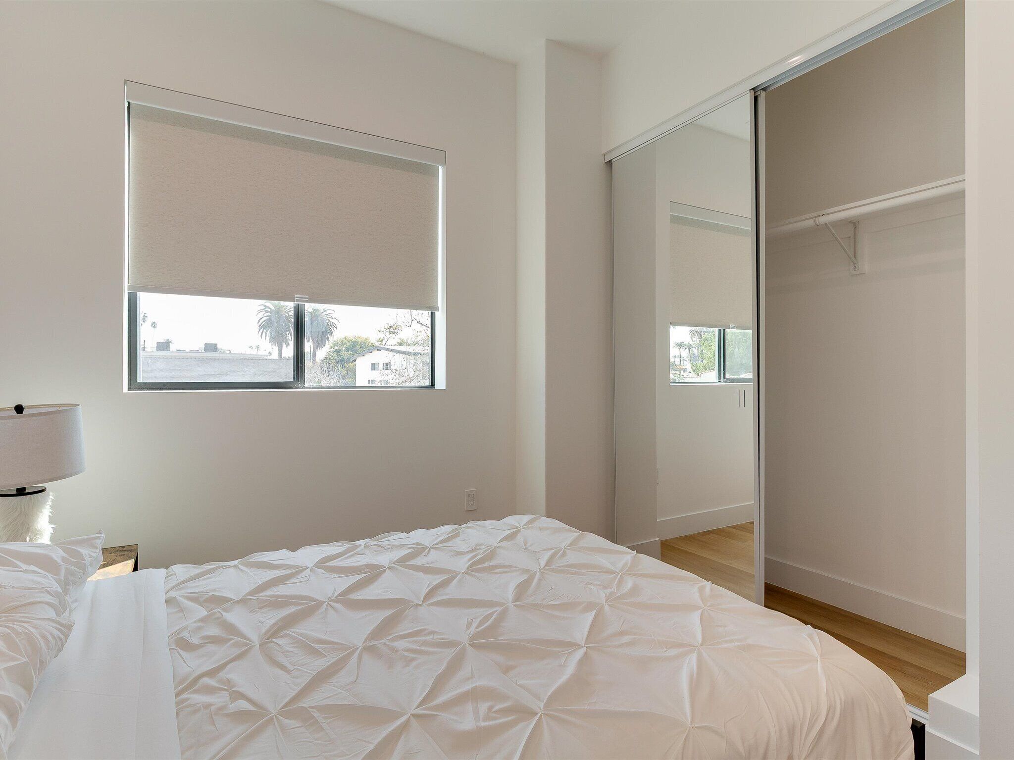 A bedroom with a bed, a window with blinds, a mirror, and a wardrobe.