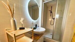 Un baño moderno con un tocador de madera, un lavabo blanco, un espejo, cabina de ducha y un florero con plumas de pampa.