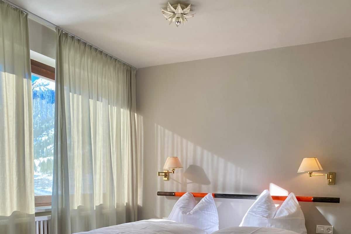 Un lit bien fait, avec du linge de lit blanc, dans une chambre d’hôtel dotée d’un sol recouvert de moquette, d’une fenêtre avec des rideaux et de deux lampes fixées au mur.