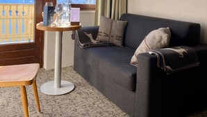 A cozy living room with a dark grey sofa, a small round table with glasses and a menu, a wooden bench, and a painting on the wall. - Arlberg Motel (Klösterle am Arlberg)