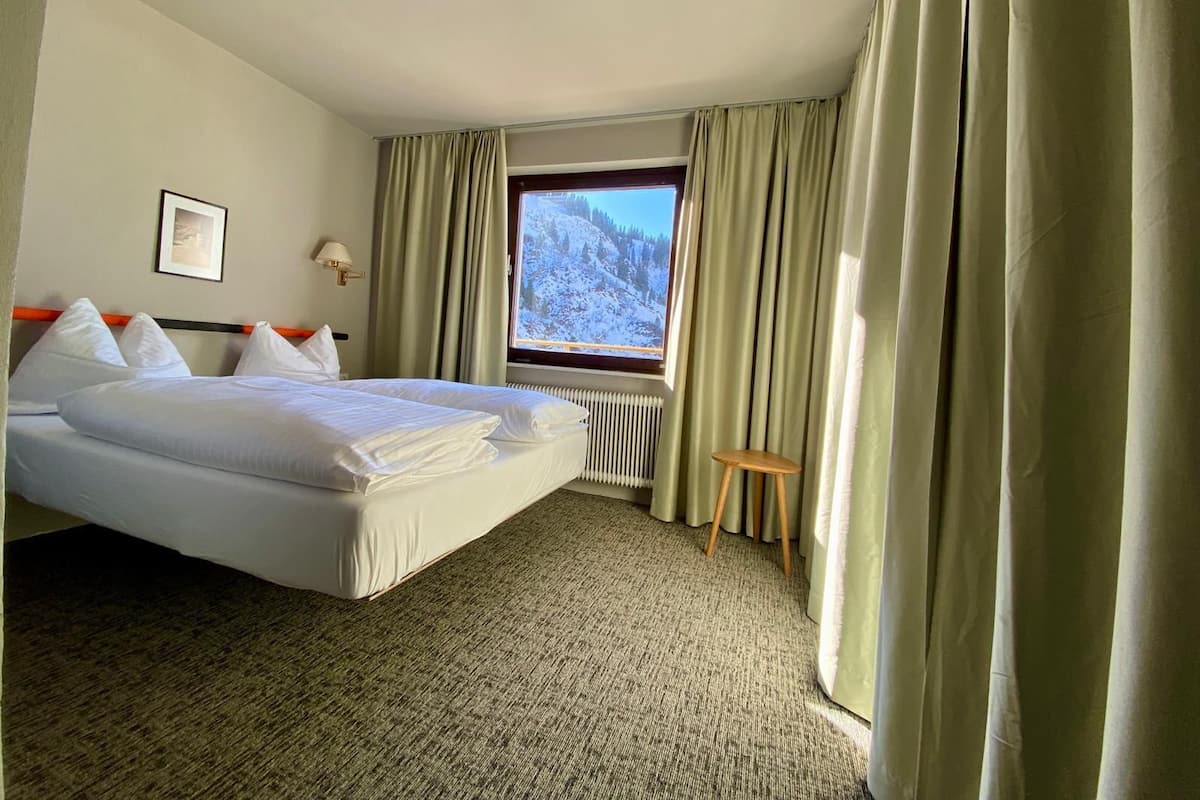 Une chambre d’hôtel avec un lit, une fenêtre donnant sur des montagnes enneigées, un radiateur et un petit tabouret.