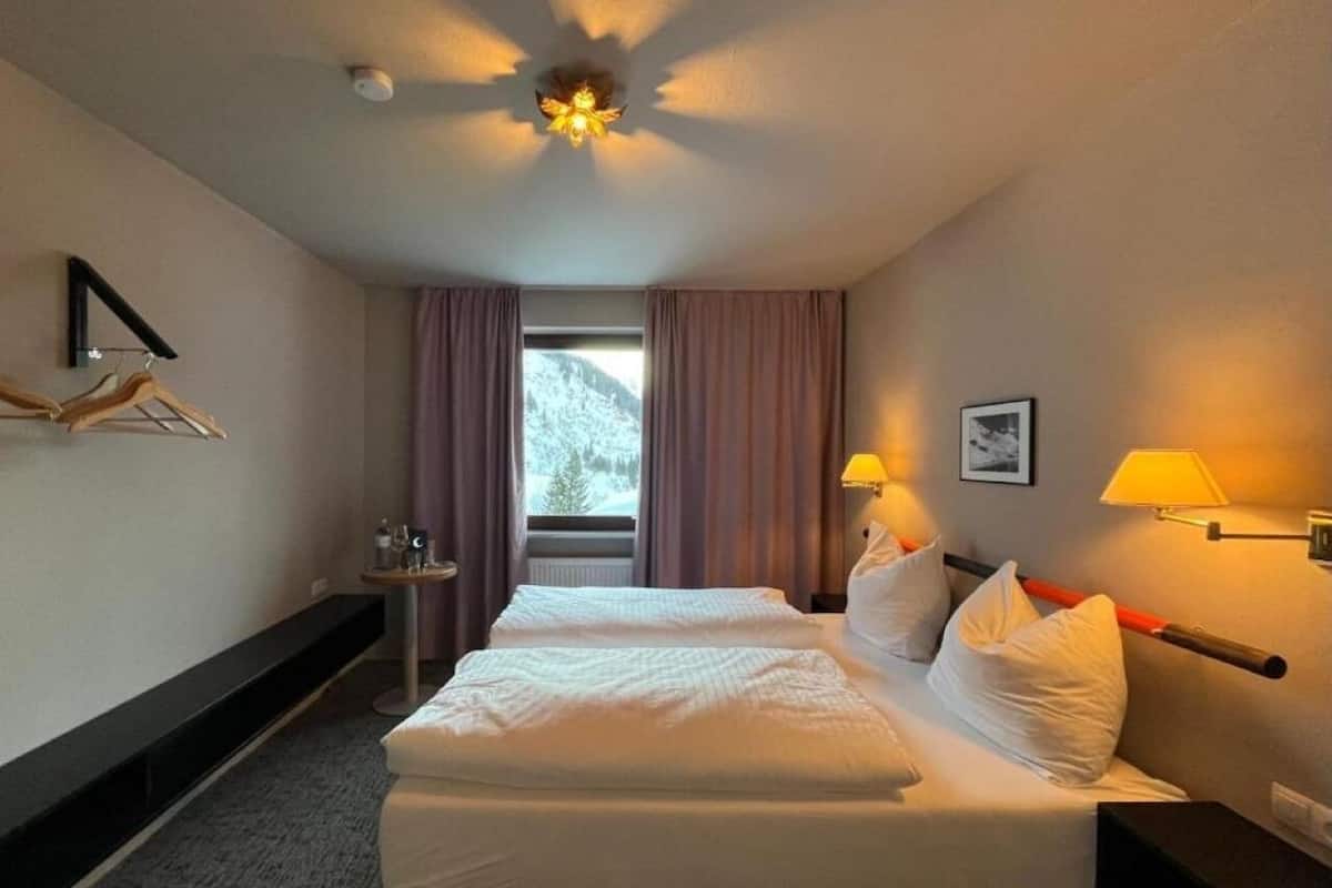 Une chambre d’hôtel avec un lit, deux lampes de chevet, une fenêtre avec des rideaux, une petite table avec une lampe et une étagère fixée au mur.
