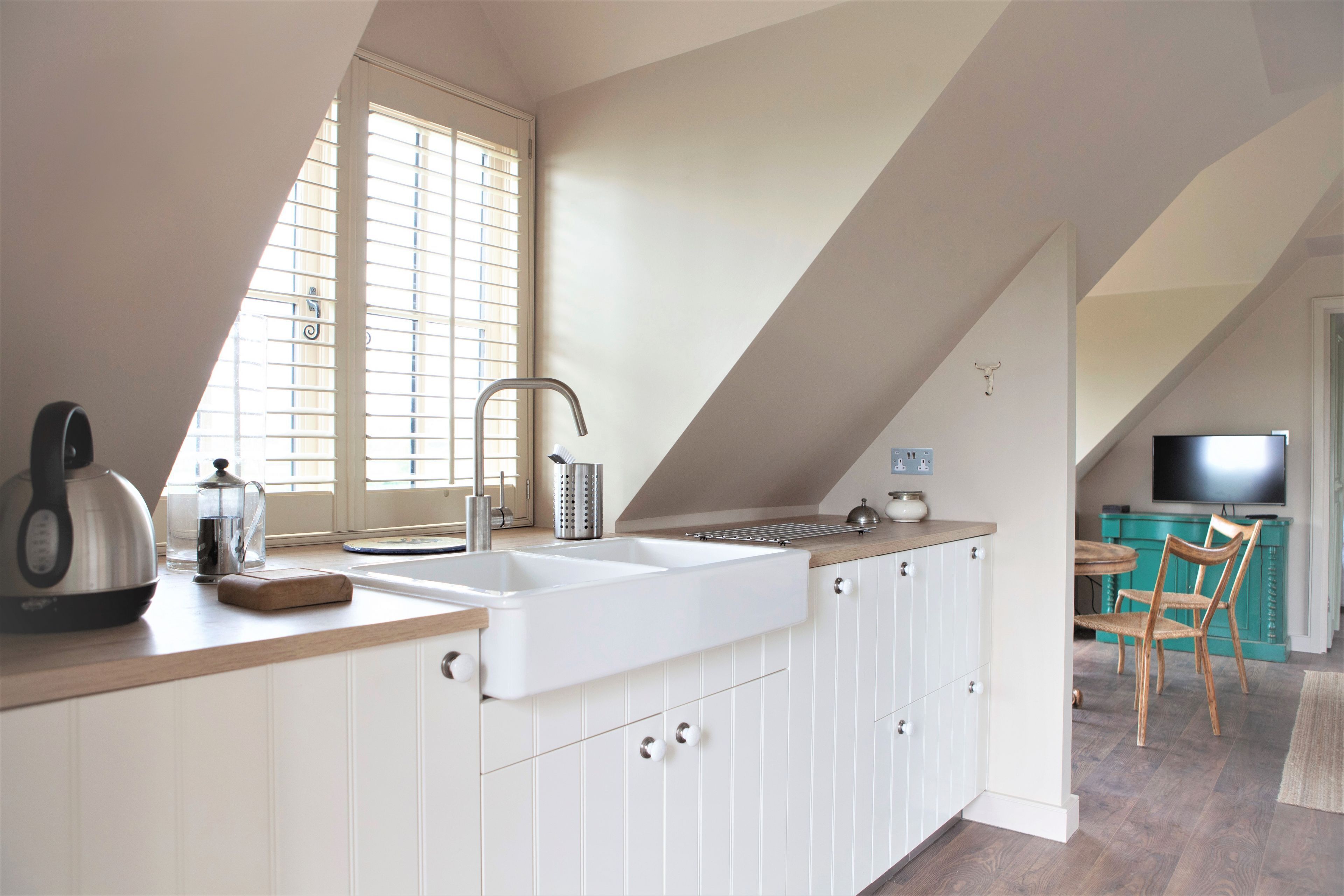 A kitchen with a large white sink, a kettle, and a coffee maker on the counter. There is a window with blinds and a television in the background.