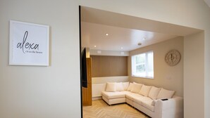 A modern living room with a white sectional sofa, a wooden floor, a wall-mounted clock, and a framed sign that reads "Alexa clean the house". - Bath Bliss Retreat - 7 Bedroom House (Bath)