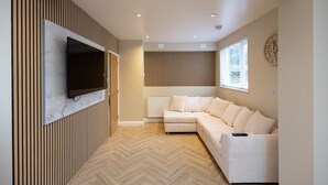A modern living room with a flat-screen TV mounted on a wooden panel wall, a white sectional sofa, and a large window with blinds. - Bath Bliss Retreat - 7 Bedroom House (Bath)