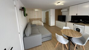 A modern living room with a grey sofa, a dining table with chairs, a kitchen area with a washing machine, and a TV. - Sublime, cosy and functional studio between braga and gêres (Bico)