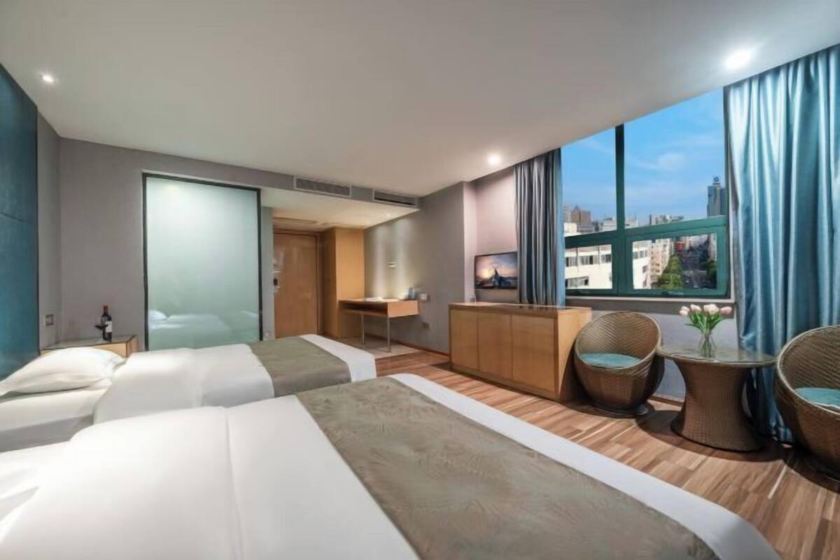 Une chambre d’hôtel avec deux lits, un bureau, une petite kitchenette, des chaises en osier et une grande fenêtre donnant sur la ville.