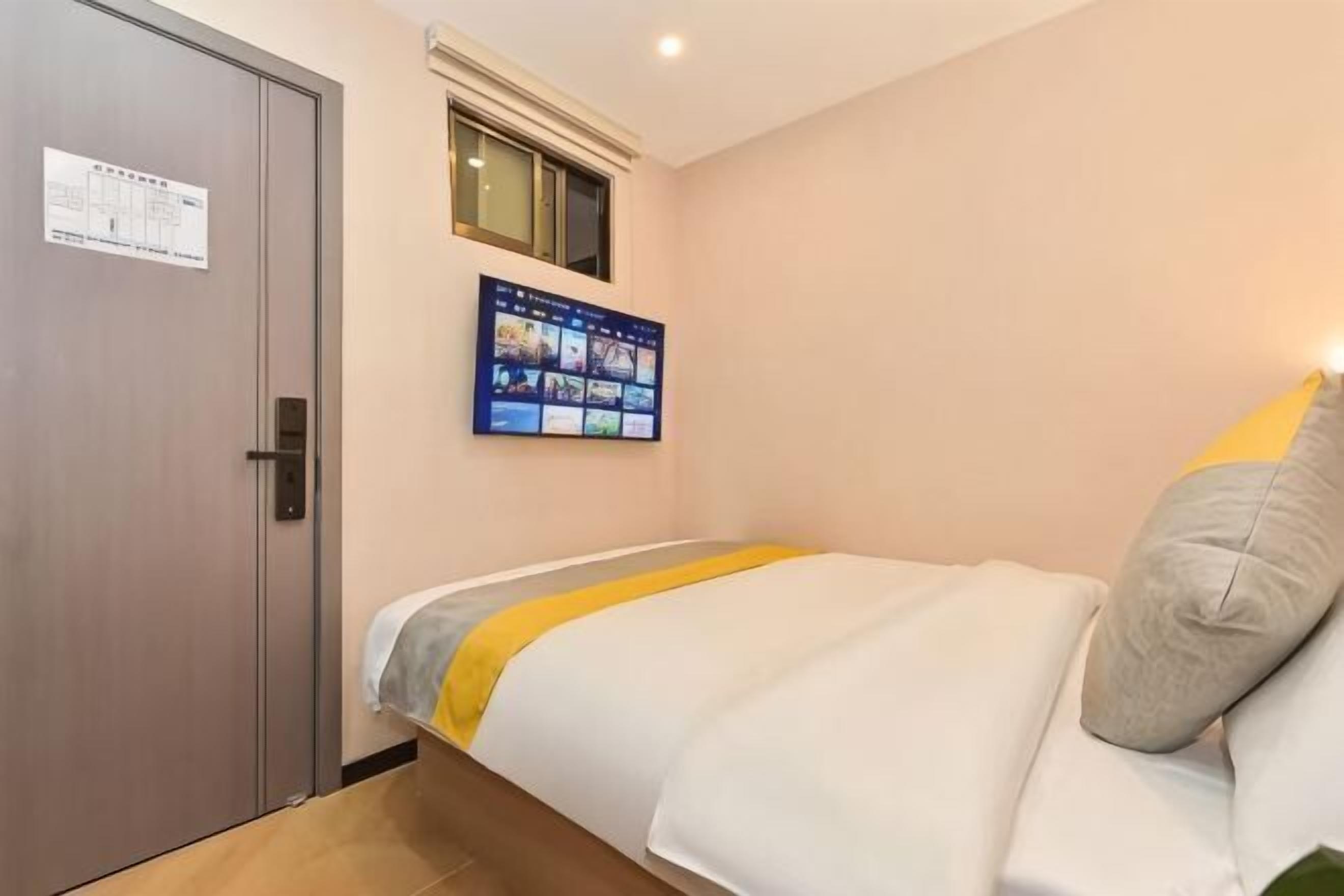 A hotel room with a bed, a door, a window, and a wall-mounted digital display.