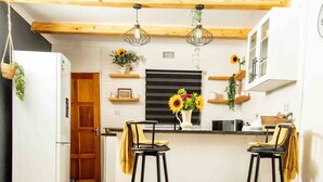 Une cuisine avec deux tabourets de bar noirs, un plafond en bois, des luminaires suspendus et un îlot central agrémenté d’un arrangement de tournesols.